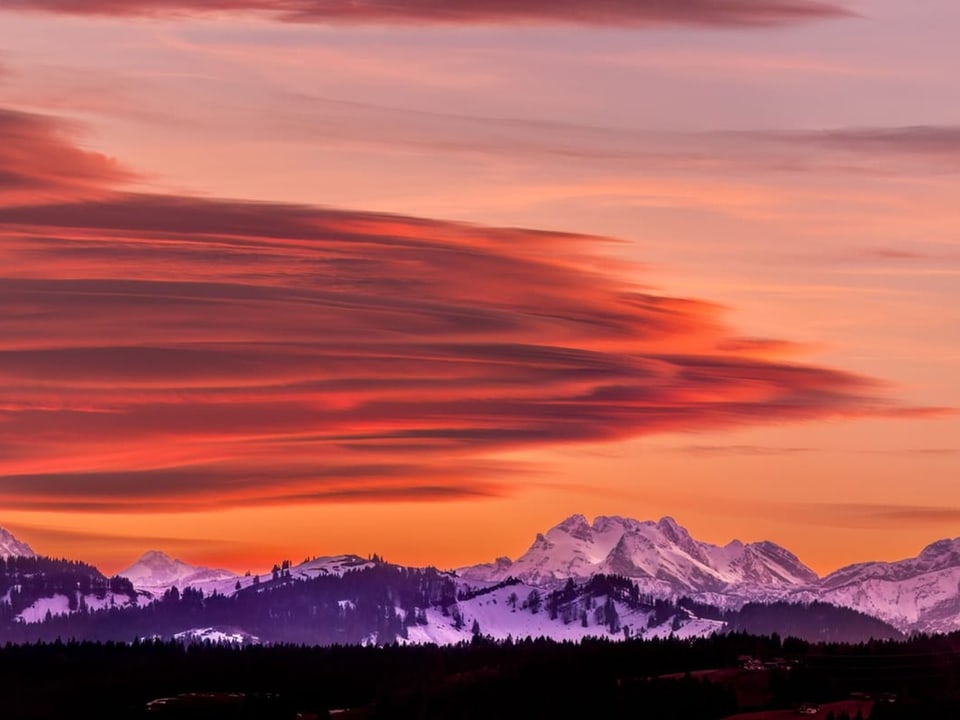 Linsenförmige rot leuchtende Wolken über verschneiten Alpen mit dunklen Waldflächen. 