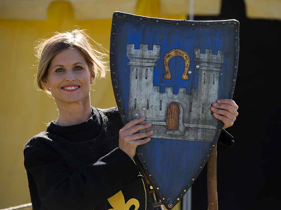 Frau in mittelalterlichem Kleid hält einen Schild hoch, das Wappen ist ein anderes als dasjenige auf ihrem Umhang