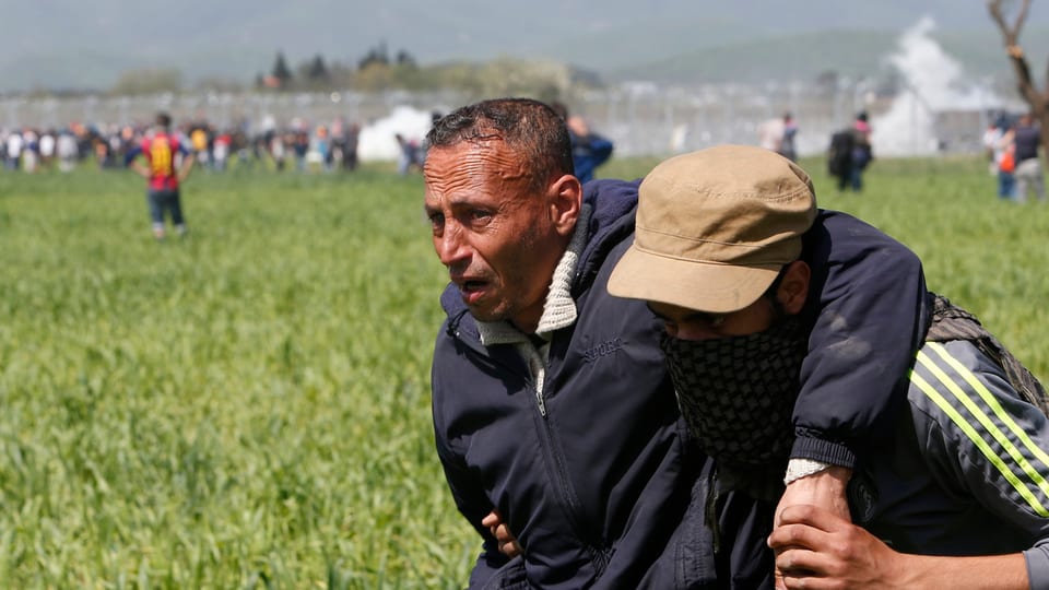 Ein sichtlich angeschlagener, älterer Mann wird von einem jüngeren Flüchtling vom Grenzzaun weggebracht.