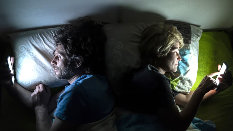 Ein Mann (links) und eine Frau liegen im Bett. Sie sind voneinander weggedreht und schauen ins helle Handy, sonst dunkel