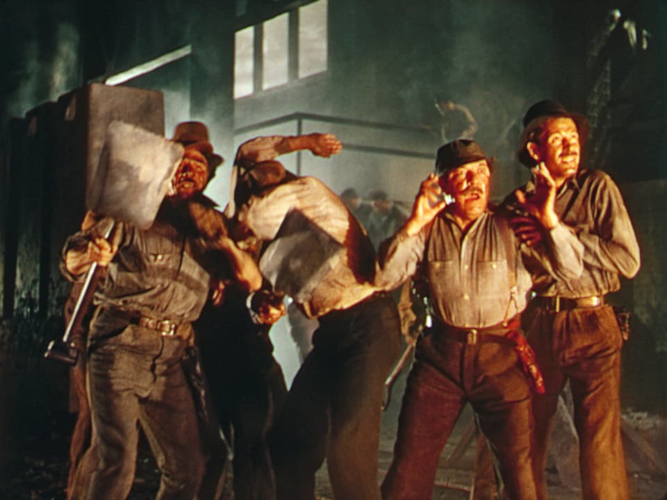 Vier Männer in einer Fabrik, deren Gesichter von einem Brand erleuchtet werden und die sich zum Teil schützend abwenden.