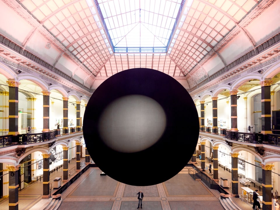 In einer klassischen Halle mit Säulen hängt eine schwarze Kugel, in der sich eine hellere Kugel befindet.