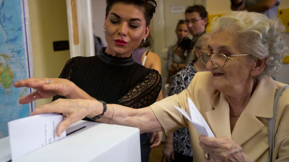Eine ältere Frau wirft in Zagreb einen Wahlzettel in die Urne, eine jüngere Angestellte hilft ihr dabei. 