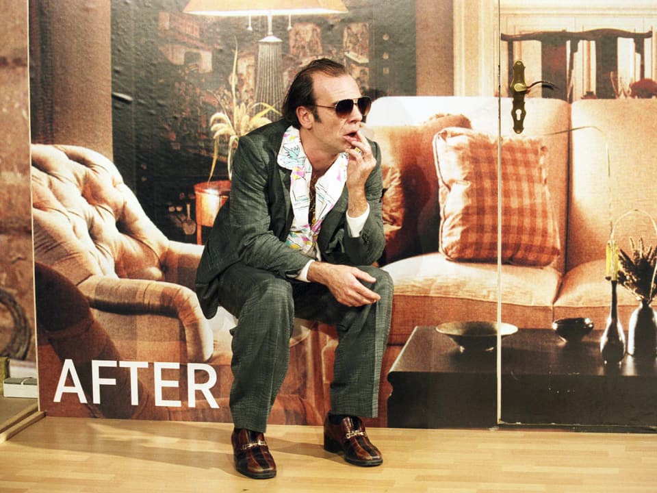 Ein Mann im Anzug und Sonnenbrille vor einer Wand mit Sofa darauf.