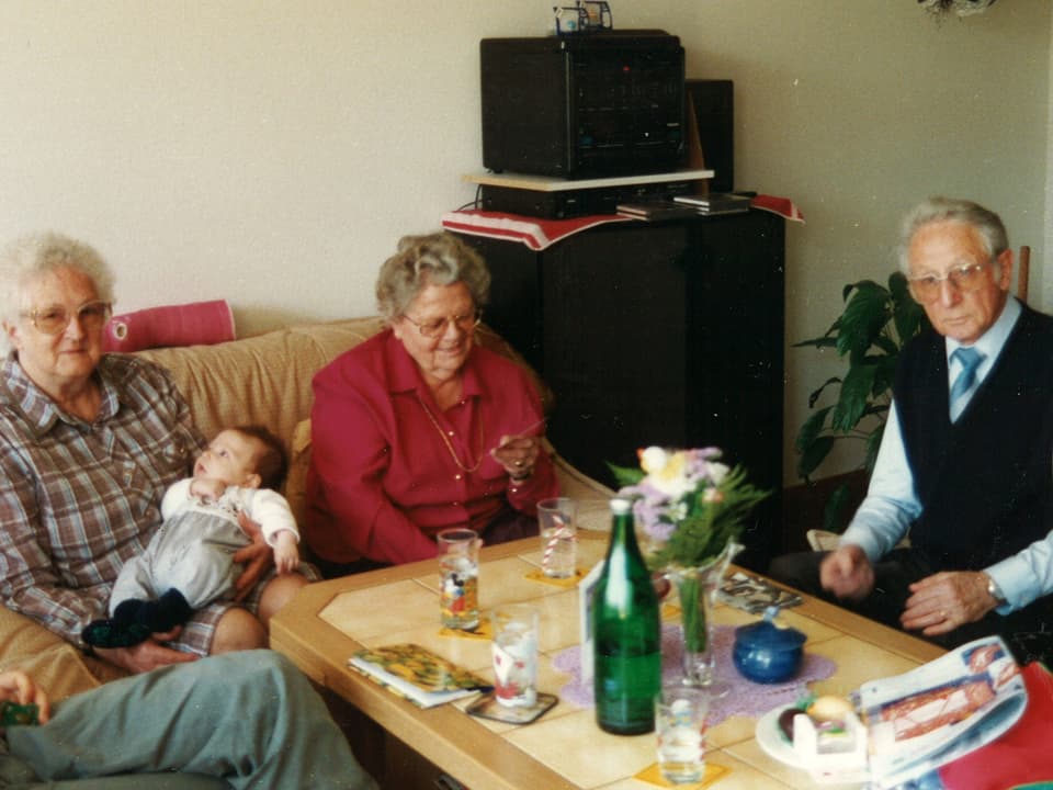 Drei ältere Senioren mit Baby an Wohnzimmertisch.