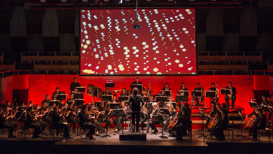 Ein Sinfonieorchester vor einer Leinwand, auf der Punkte zu sehen sind. 