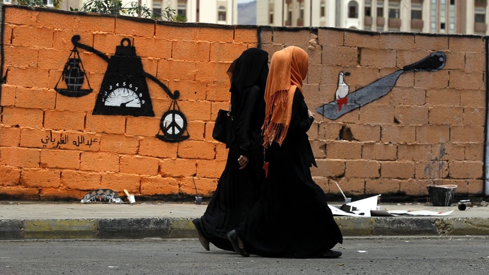 Zwei Frauen gehen an einer Mauer mit einem Graffiti entlang, darauf eine Waage mit einer Bombe und einem Friedenszeichen.