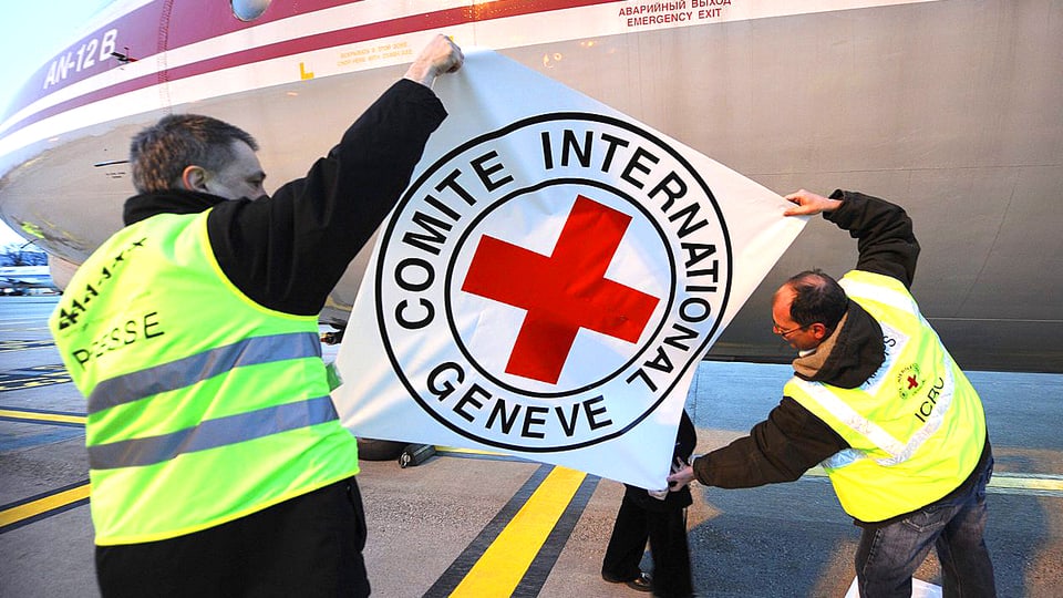 Zwei IKRK-Mitarbeiter kleben eine Folie mit dem Organisations-Emblem auf ein Transportflugzeug.