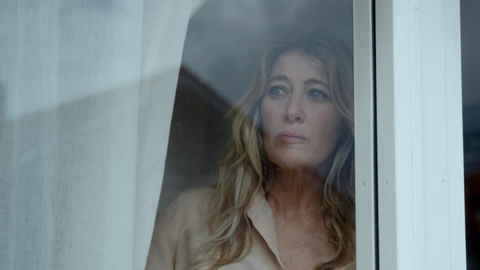 Hinter Fensterscheibe schaut blonde Frau mit traurigem Blick aus dem Fenster
