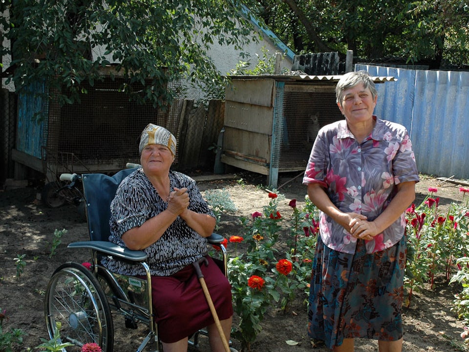 Eine alte Frau im Rollstuhl sitzt an der Seite ihrer Schwester im Garten mit roten Blumen.