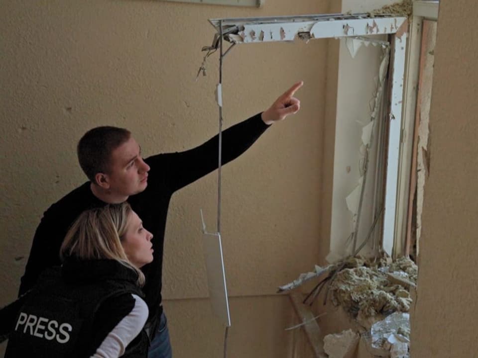  Luzia Tschirky trägt eine Medien-Veste. Sie steht neben einem Mann, der aus einem kaputten Fenster zeigt.