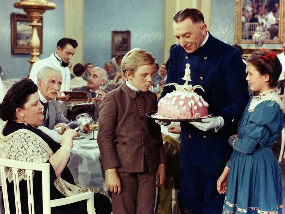Ein Mann hält einen grossen Kuchen vor sich. Links und rechts davon stehen Peter und Heidi die den Kuchen mit grossen Augen betrachten.