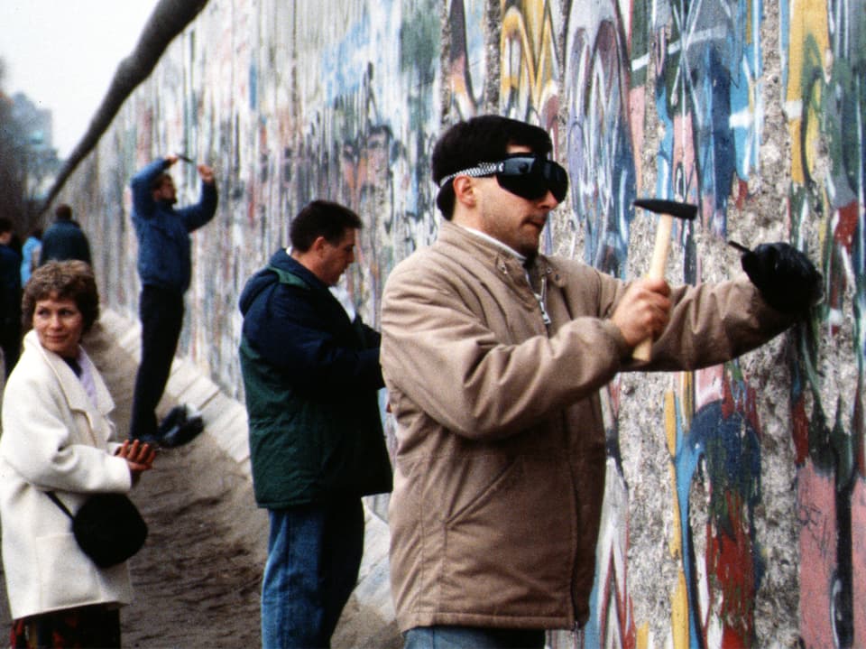 Mehrere Menschen hauen mit Hammern in die Berliner Mauer.