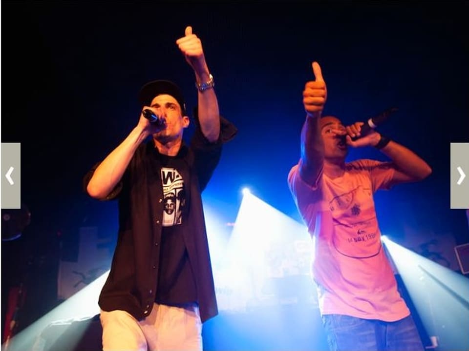 Zu sehen sind zwei Rapper auf der Bühne. Sie halten ein Mikrofon vor ihren Mund und heben den Daumen der anderen Hand.