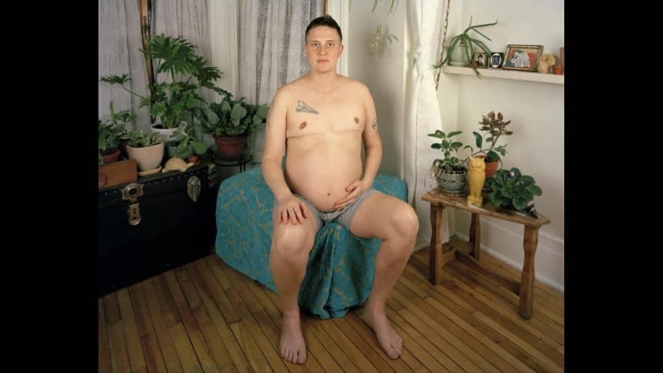 Ein Mann mit nacktem Oberkörper und dickem Schwangerschaftsbauch sitzt in einem Wohnzimmer