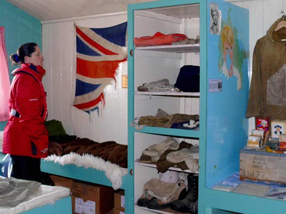 Eine Frau steht in einem kabinenartigen Raum mit Pritsche und Regal. An der Wand hängt eine zerrissene Grossbritannien-Fahne. 
