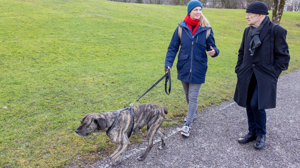 Eine Frau mit Kappe hält einen Hund an der Leine und spricht mit einem Mann, der neben ihr läuft.
