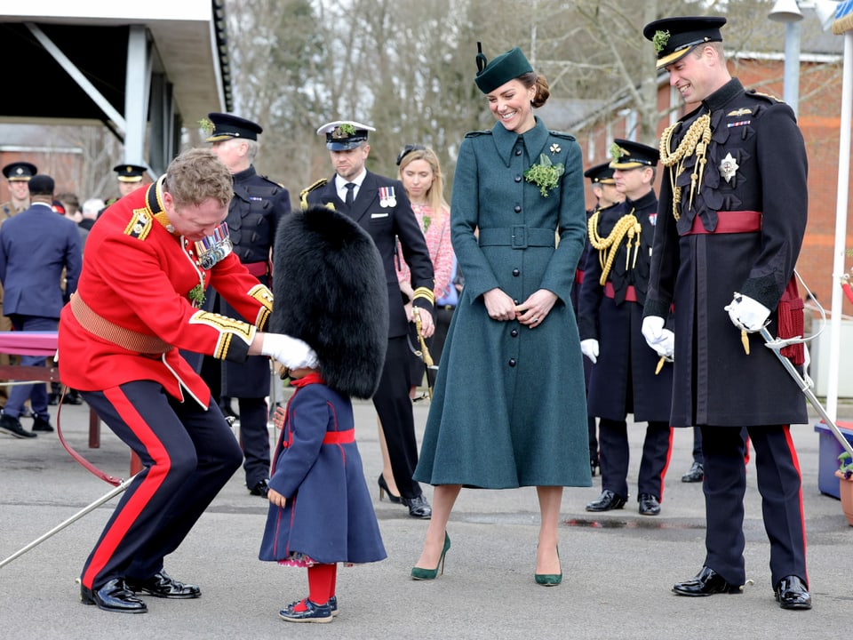 Ein Mann in Uniform setzt seiner kleinen Tochter die riesige Bärenmütze auf. Herzogin Kate und Prinz William stehen lachend daneben.