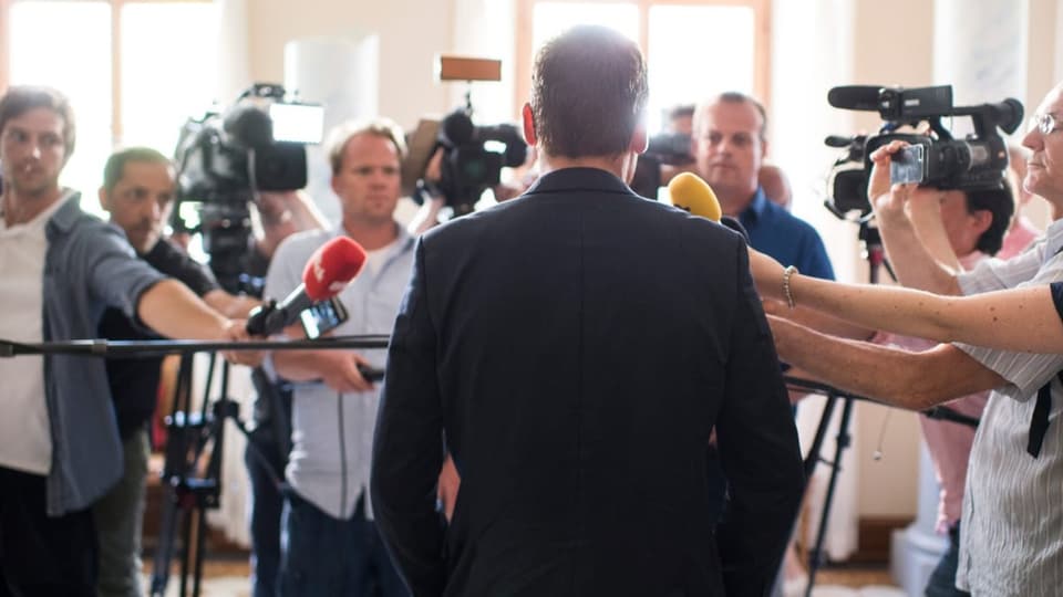 Mann mit Rücken zur Bild, im Hintergrund Reporter mit Kameras und Mikrofonen zu ihm hingestreckt.