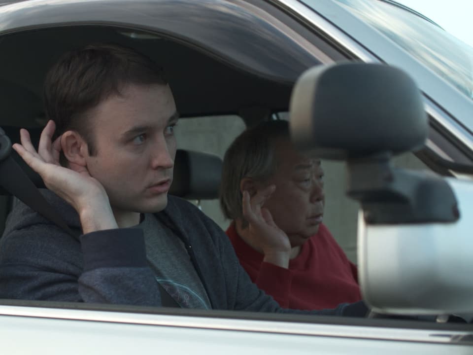 Zwei Männer in einem Auto, die eine Hand an ihr Ohr halten.