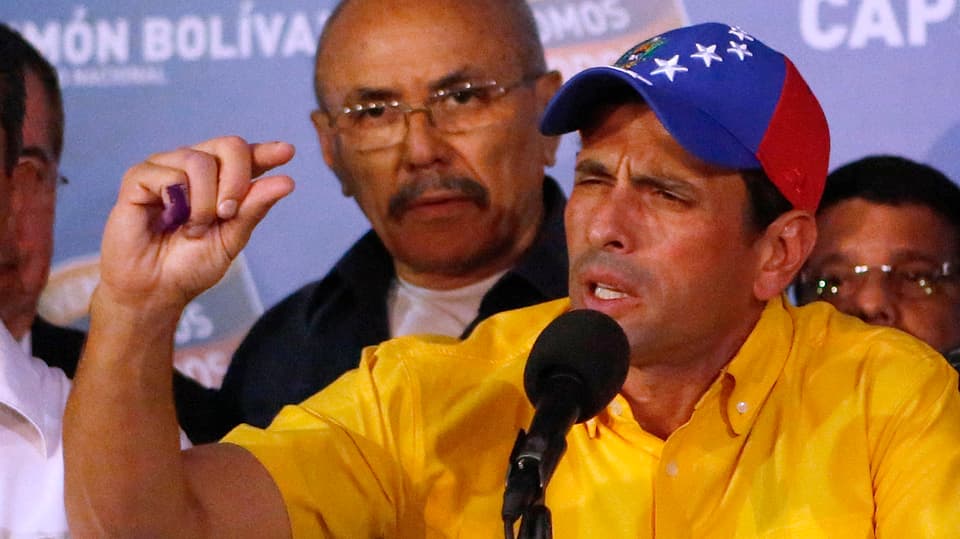 Capriles im gelben Shirt deutet mit Daumen und Zeigefinger an einer Pressekonferenz den kleinen Abstand zwischen den Resultaten an.