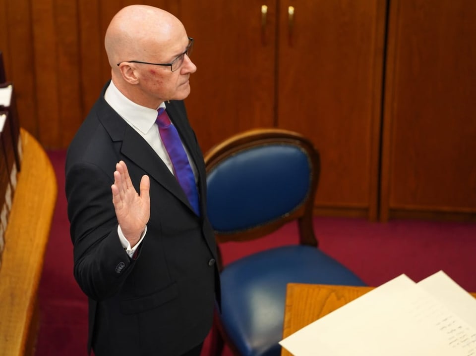 Glatzköpfiger Mann in Anzug leistet Schwur neben Dokument auf Schreibtisch im Gerichtssaal.