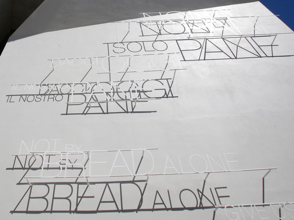 Graue Wand mit Aufschrift «nicht vom Brot allein» in verschiedenen Sprachen