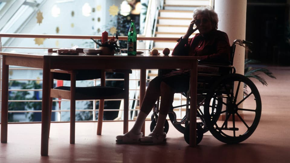 gegenlichtaufnahme einer Seniorin im Rollstuhl, die alleine an einem Tisch sitzt und den Kopf aufstützt.