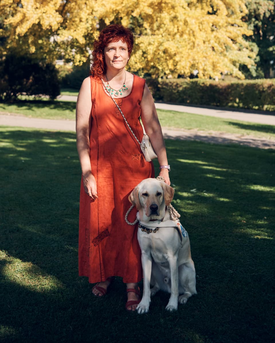 Eine Frau in rotem Jumpsuit mit lockigen Haaren hält rechts einen Stock, links das Hundegeschirr. Labrador geht vor ihr.