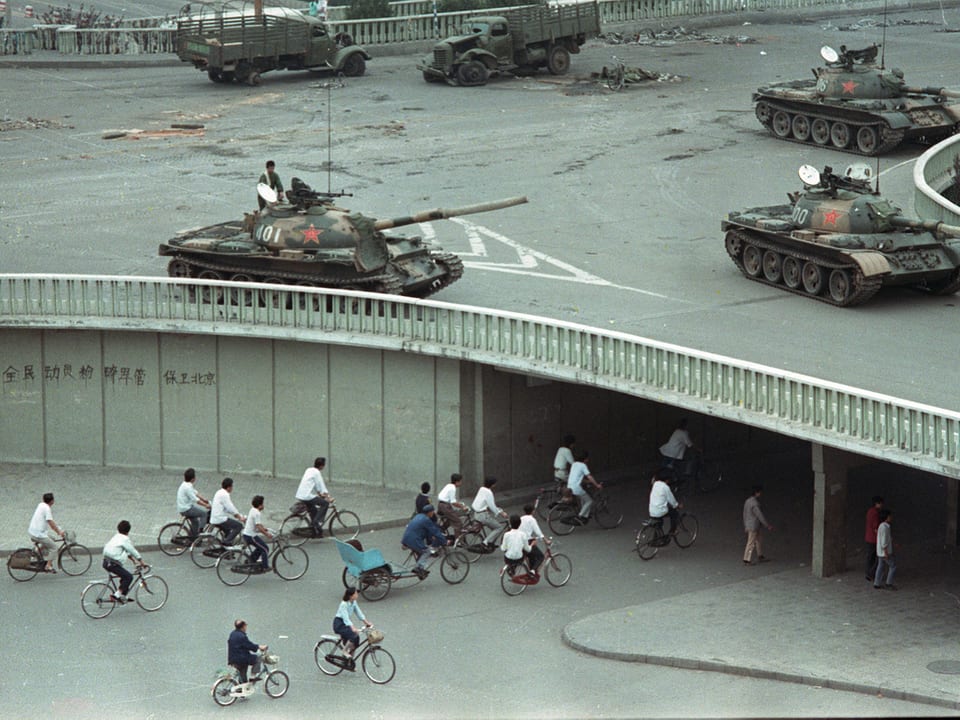 Velofahrer in Peking fahren unter einer Kreuzung durch. Auf dieser Kreuzung stehen Panzer der Armee.