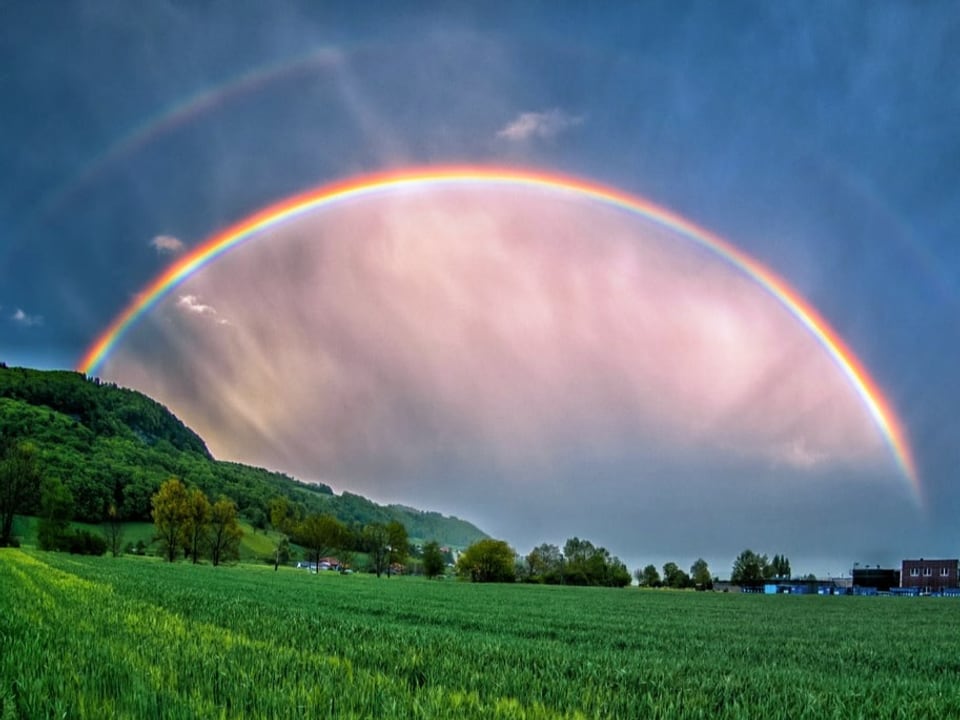 Dunkle Wolken, vor denen ein Regenbogen mit Doppelbogen leuchtet. Unten satt grünes Feld. 