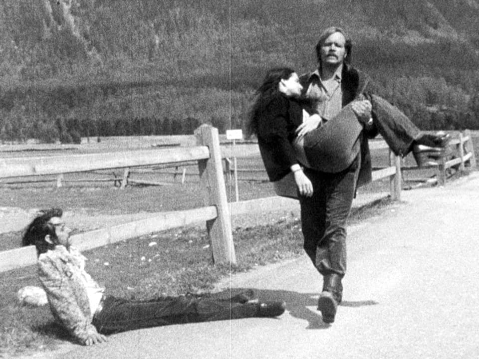 Ein Mann trägt eine ohnmächtige Frau eine Strasse entlang, am Strassenrand lehnt ein Mann leblos an ein Geländer.