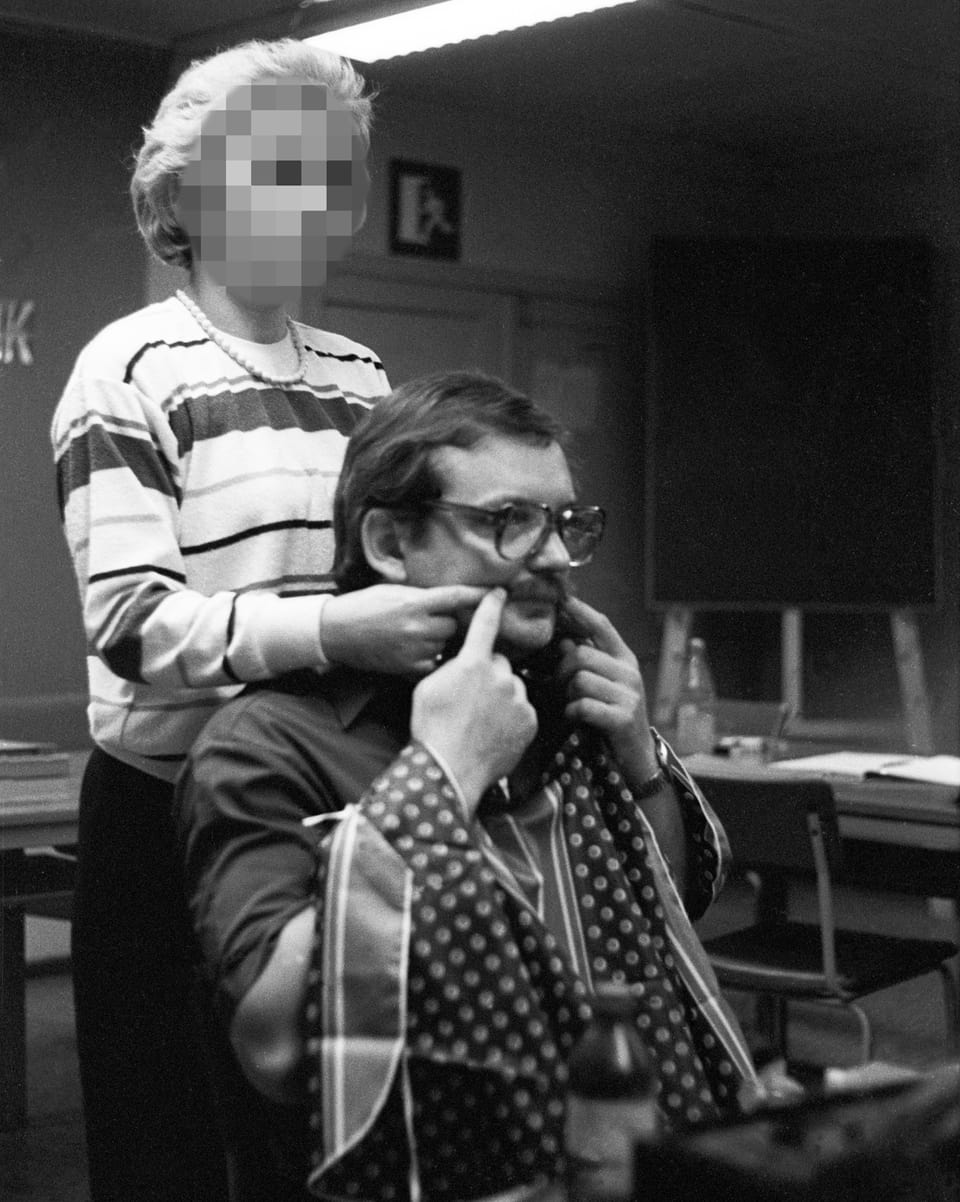 Schwarz-Weiss-Foto: Ein Mann sitz auf einem Stuhl und klebt sich einen falschen Schnauz an. Eine Frau hilft ihm dabei