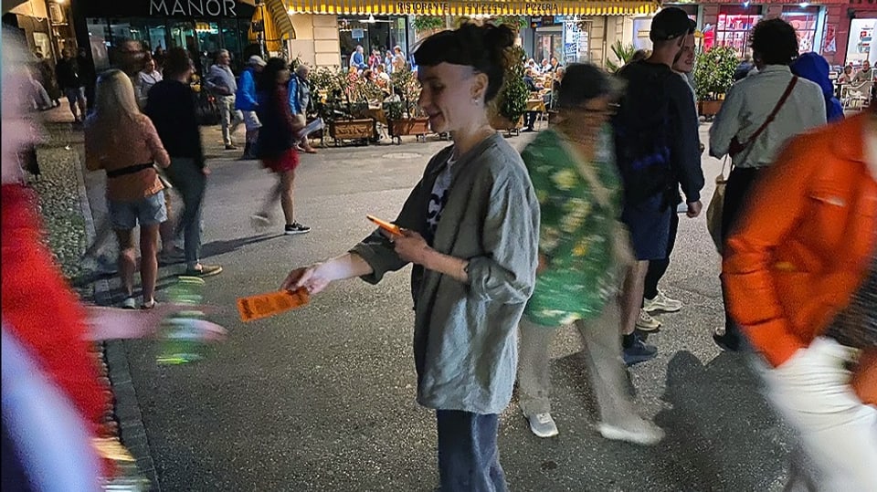 Junge Frau in grauem Hemd steht auf Asphalt, verteilt orange Flyer an verschwommene Passanten.