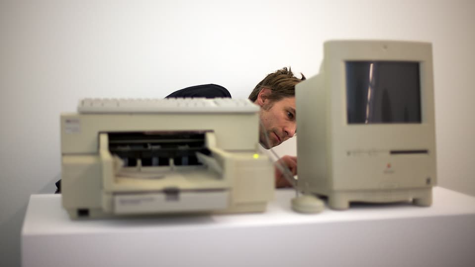 Ein Mann steht hinter einem alten Drucker und einem alten Macintosh-Computer und scheint ein Kabel umzustecken