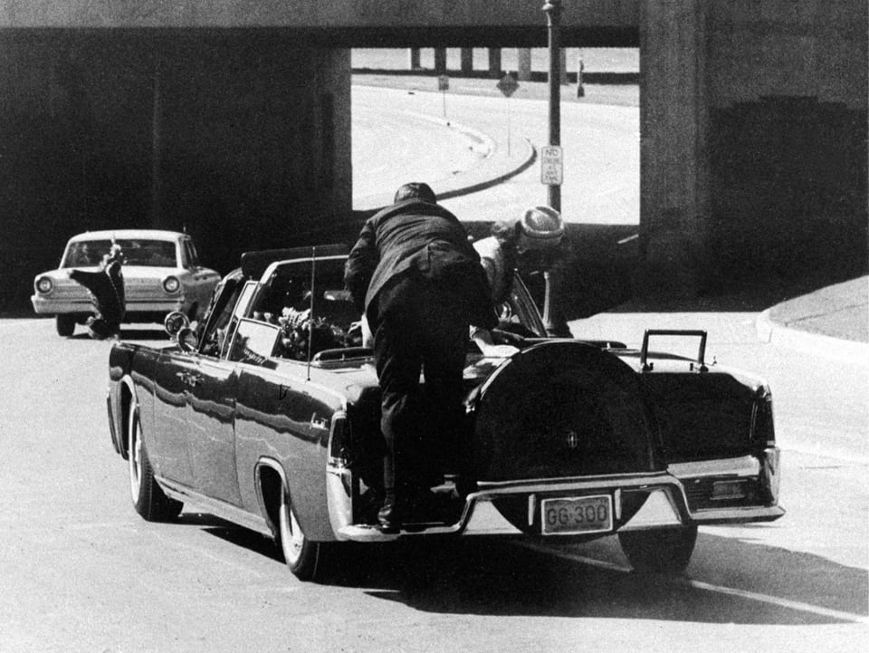 Sekunden nach dem Attentat auf JFK, ein Sicherheitsbeamter ist auf den Wagen aufgesprungen, Jackie beugt sich über den schwerverletzten Präsidenten.