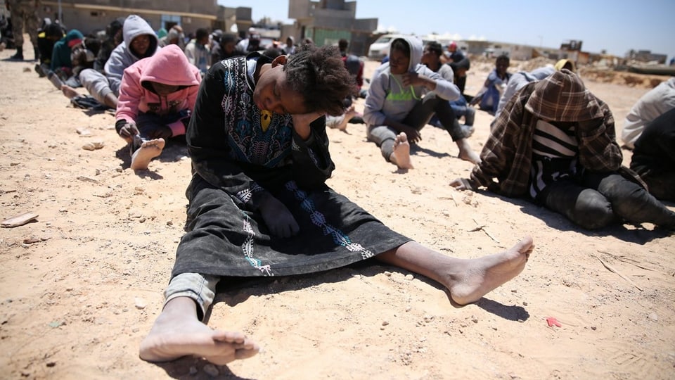 Erschöpfte Flüchtlinge sitzen auf staubigem Boden.