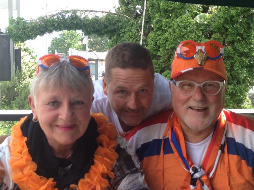 Älteres Ehepaar mit orangefarbenen Brillen, Fan-Mütze und Fan-Girlande geschmückt. Im Hintergrund ein Mann. Alle drei lachen in die Kamera.