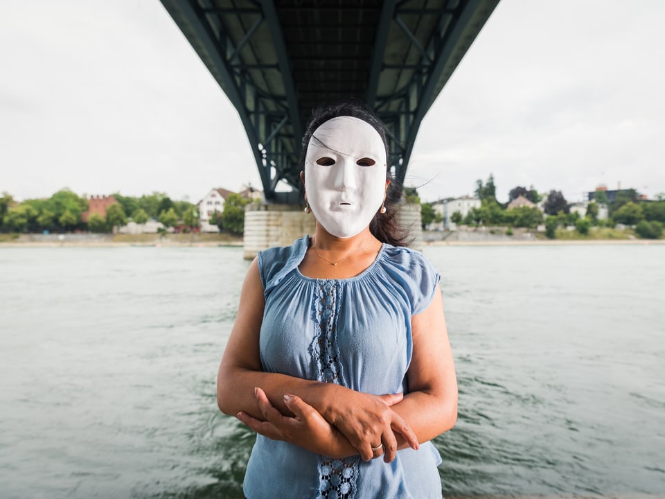 Eine Frau trägt eine weisse Maske um ihre Identität zu verbergen. Sie steht nahe des Rheins in Basel, unter einer Brücke.