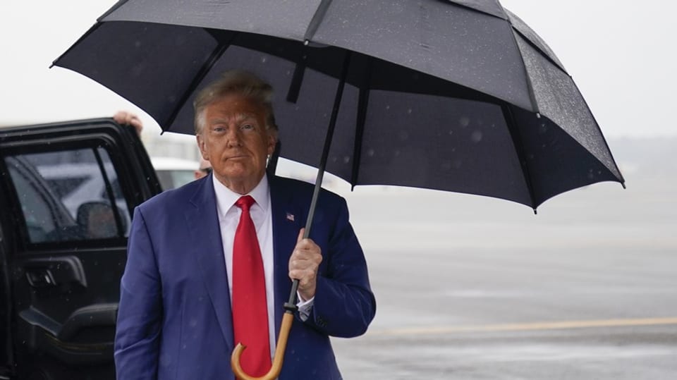 Trump mit einem Regenschirm. Im Hintergrund ist ein Auto zu sehen.