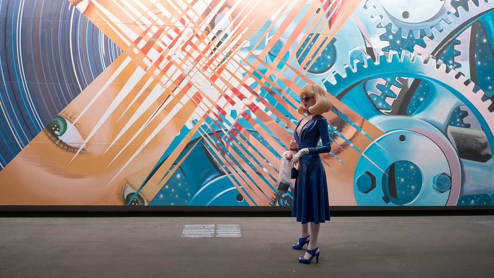 Eine Frau aus Plastik steht vor einer riesigen, collageartigen Malerei.