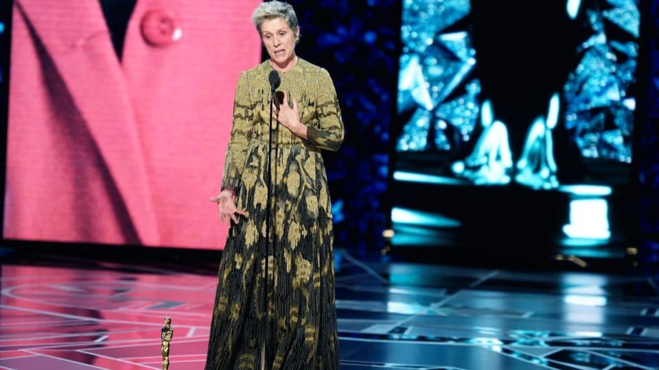 Eine Frau im langen Kleid steht auf einer Bühne. Vor ihr ein Mikrofon, neben ihr auf dem Boden eine Trophäe.