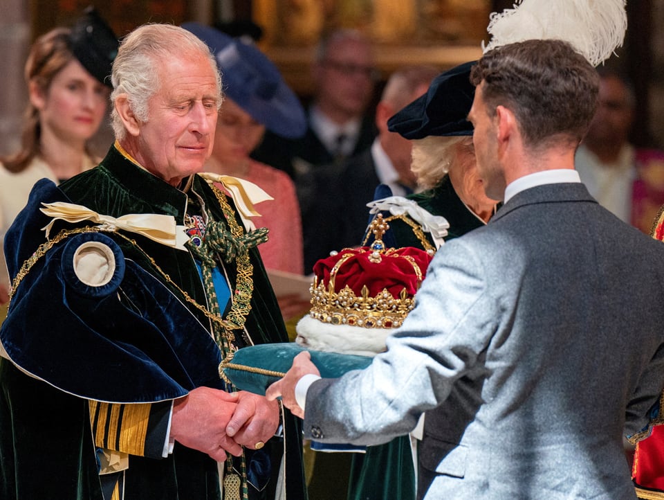 King Charles III. steht in königlichem Gewand. Ein Mann hält ihm eine Krone auf einem Kissen hin.