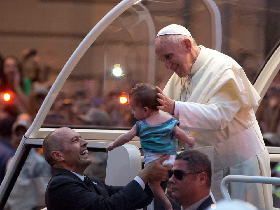 Der Papst segnet ein Kind, dass ihn von einem Sicherheitsmann gereicht wird.