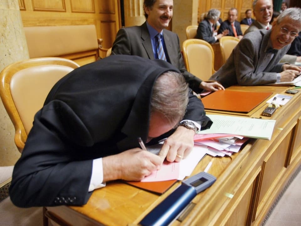 Boris Banga füllt seinen Stimmzettel aus, indem er die Jacke über den Kopf und den Zettel zieht. Seine Ratskollegen lachen. 