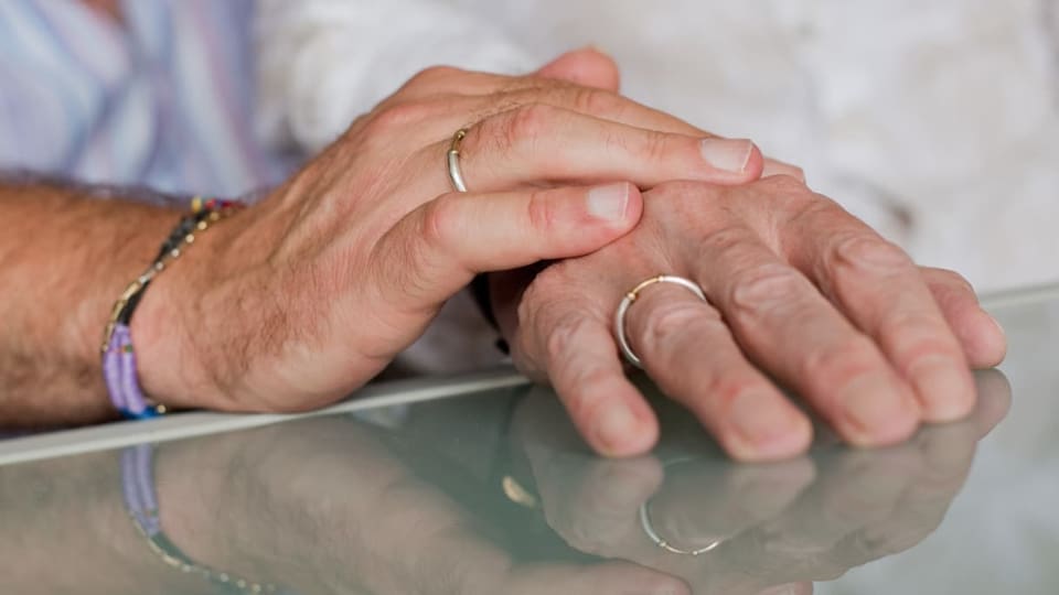 Zwei Männer tragen Eheringe und legen ihre Hände übereinander auf einem Tisch.