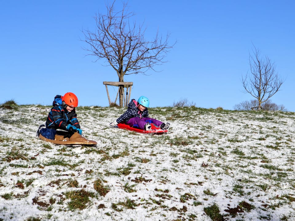 Zwei Kinder versuchen mit ihrem Bob die Abfahrt auf einer dünnen Schneedecke, teils dringt Gras durch die Schneeauflage.