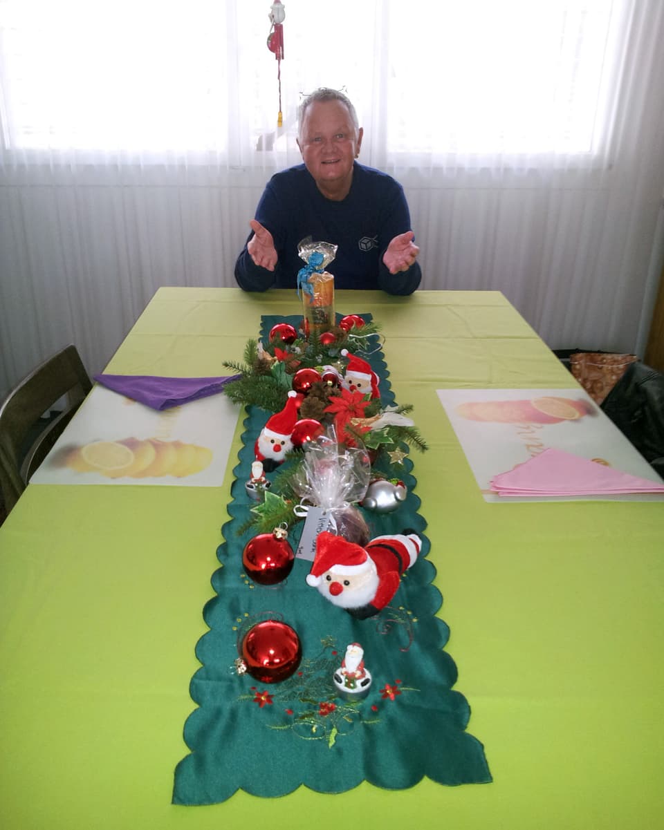 Jörg Stoller sitzt an einem langen Tisch, auf dem in der Mitte ein grüner Tischläufer liegt. Auf dem Tischläufer hat es rote Samichläuse und Weihnachtskugeln.