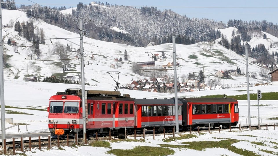 Ein roter Zug mit drei Waggons rollt durch eine Winterlandschaft.