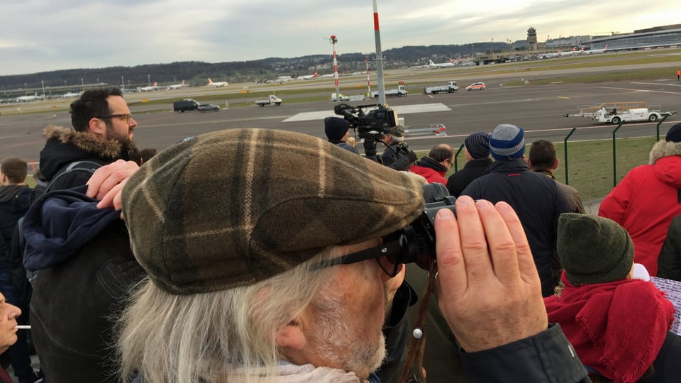 Mann mit grauen Haaren und Kappe sieht mit Feldstecher auf den Flughafen